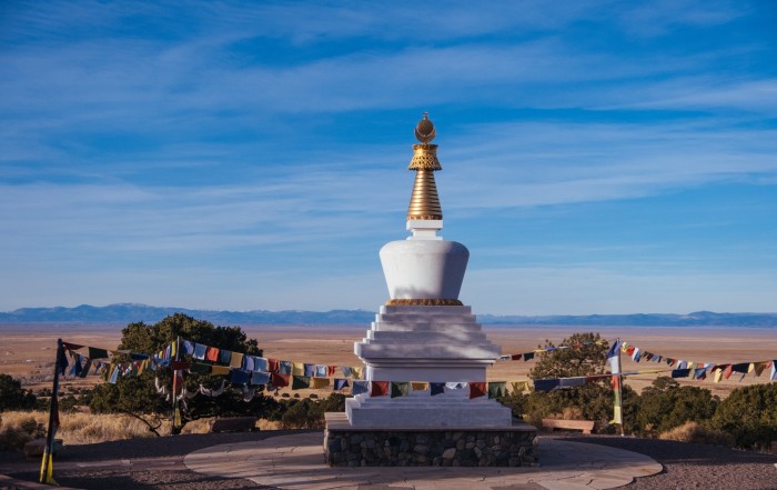 A white stupa with a gold spire under a wide blue sky, prayer flags strung across a high desert valley