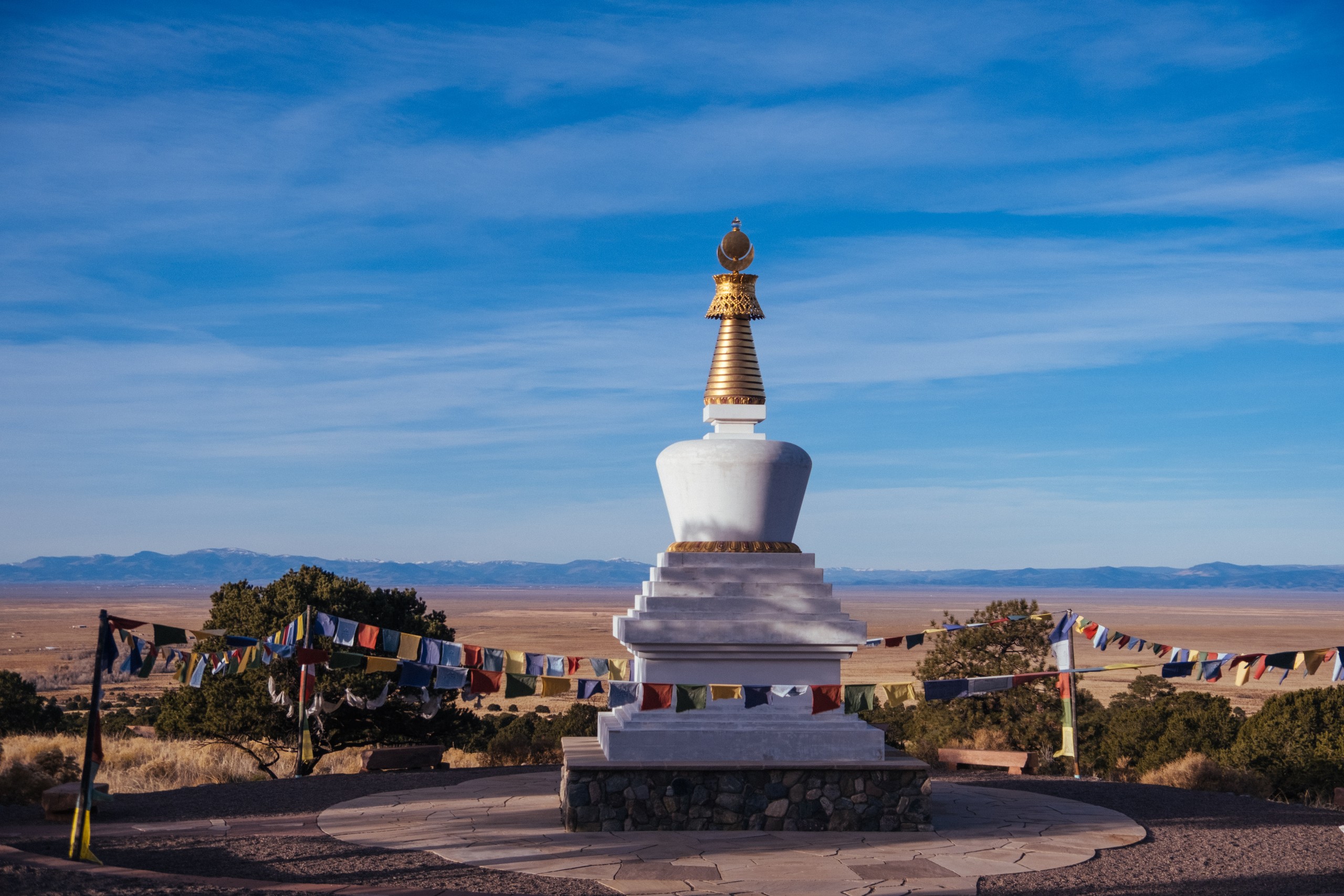 A white stupa with a gold spire under a wide blue sky, prayer flags strung across a high desert valley