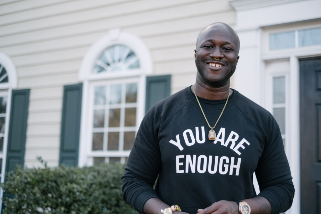 A smiling man in a 'You Are Enough' sweatshirt photographed outside his home