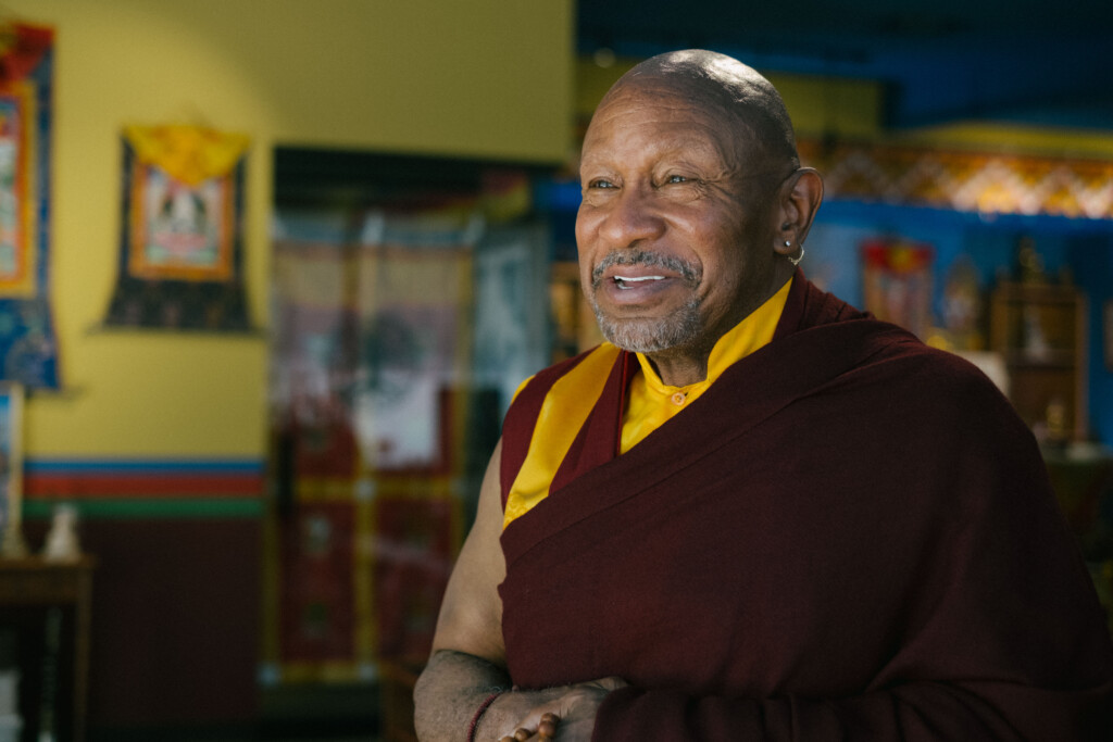 A smiling man in maroon robes photographed in a colorful shrine room
